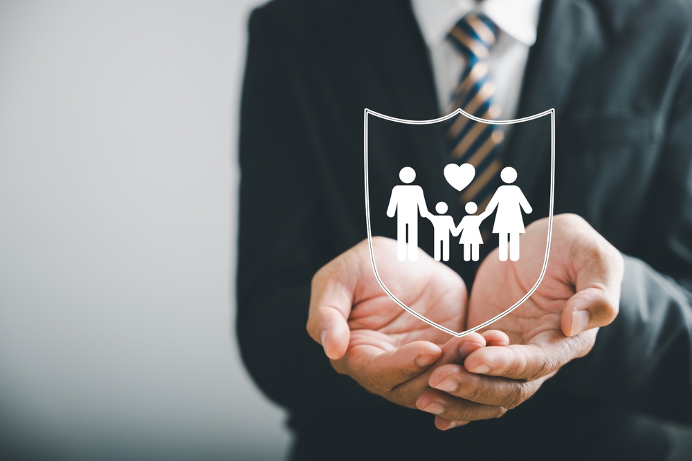 A person in a business suit holding a transparent icon of a family inside a shield, symbolizing family protection and insurance.